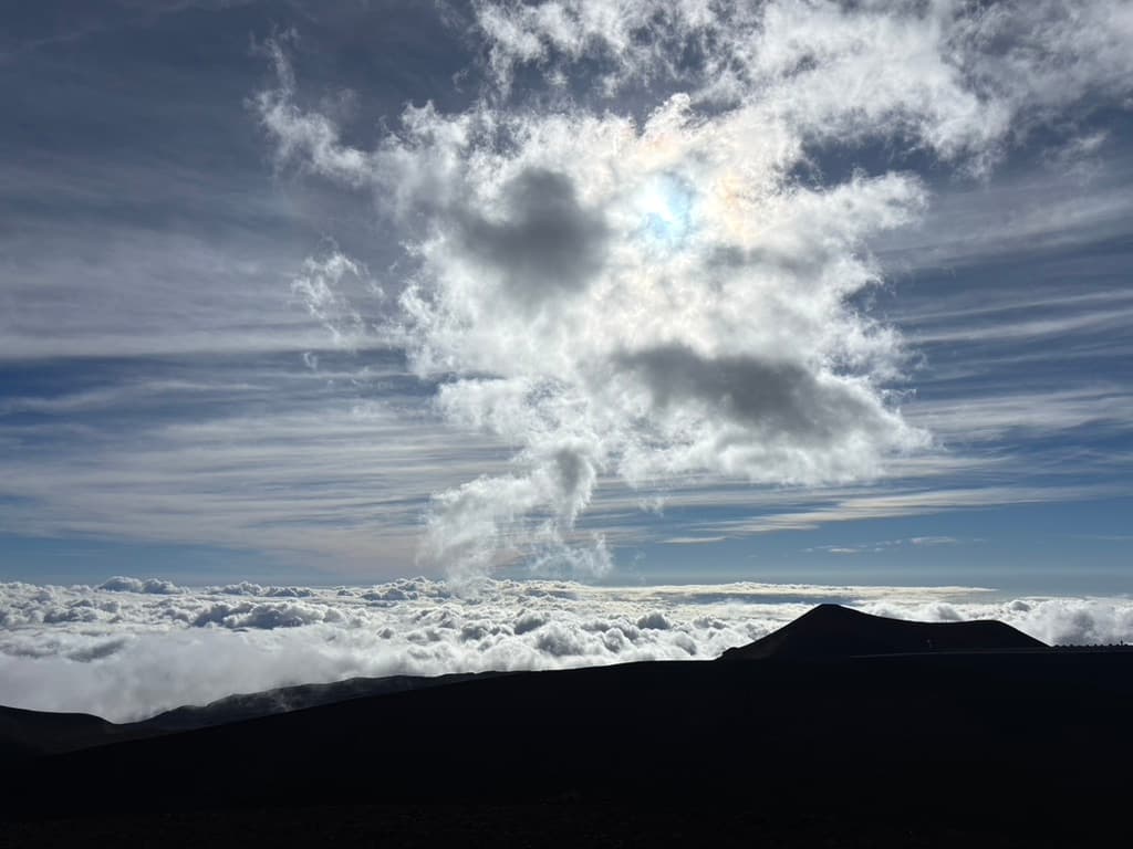 Sunlit clouds rising above a dark Mauna landscape and a broad layer of clouds