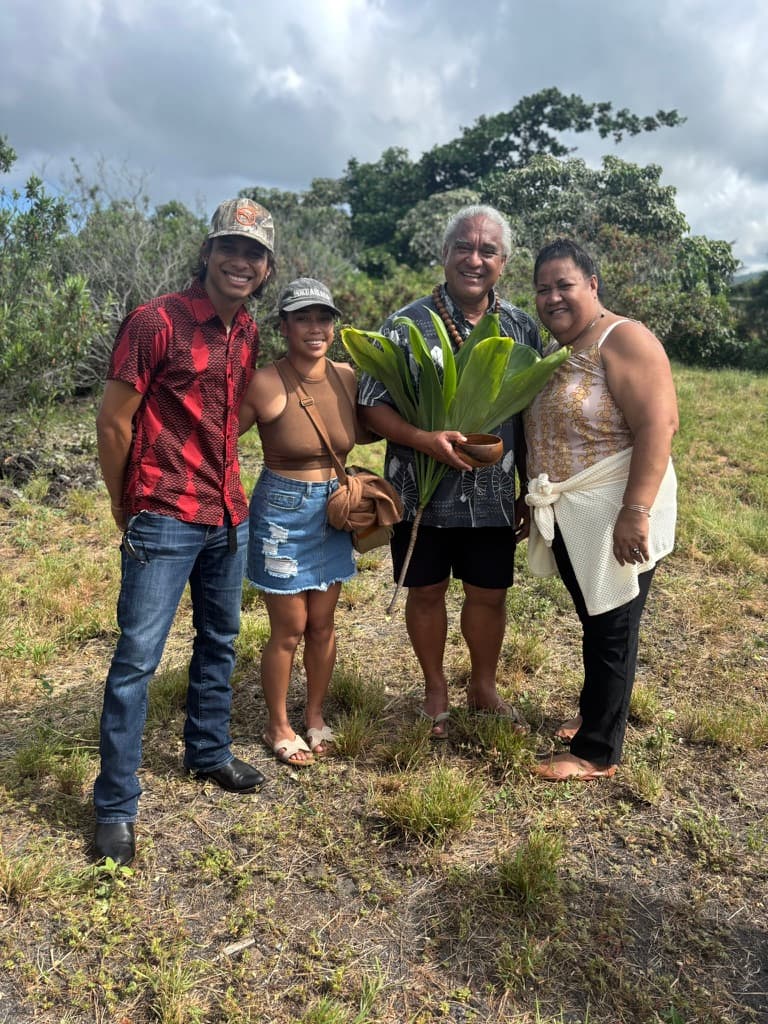 Four people standing together outdoors, one holding a bowl and green ti leaves
