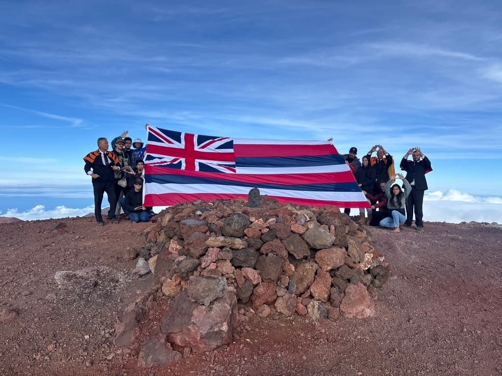 A group gathered on the Mauna holding a large Hawaiian flag above the clouds